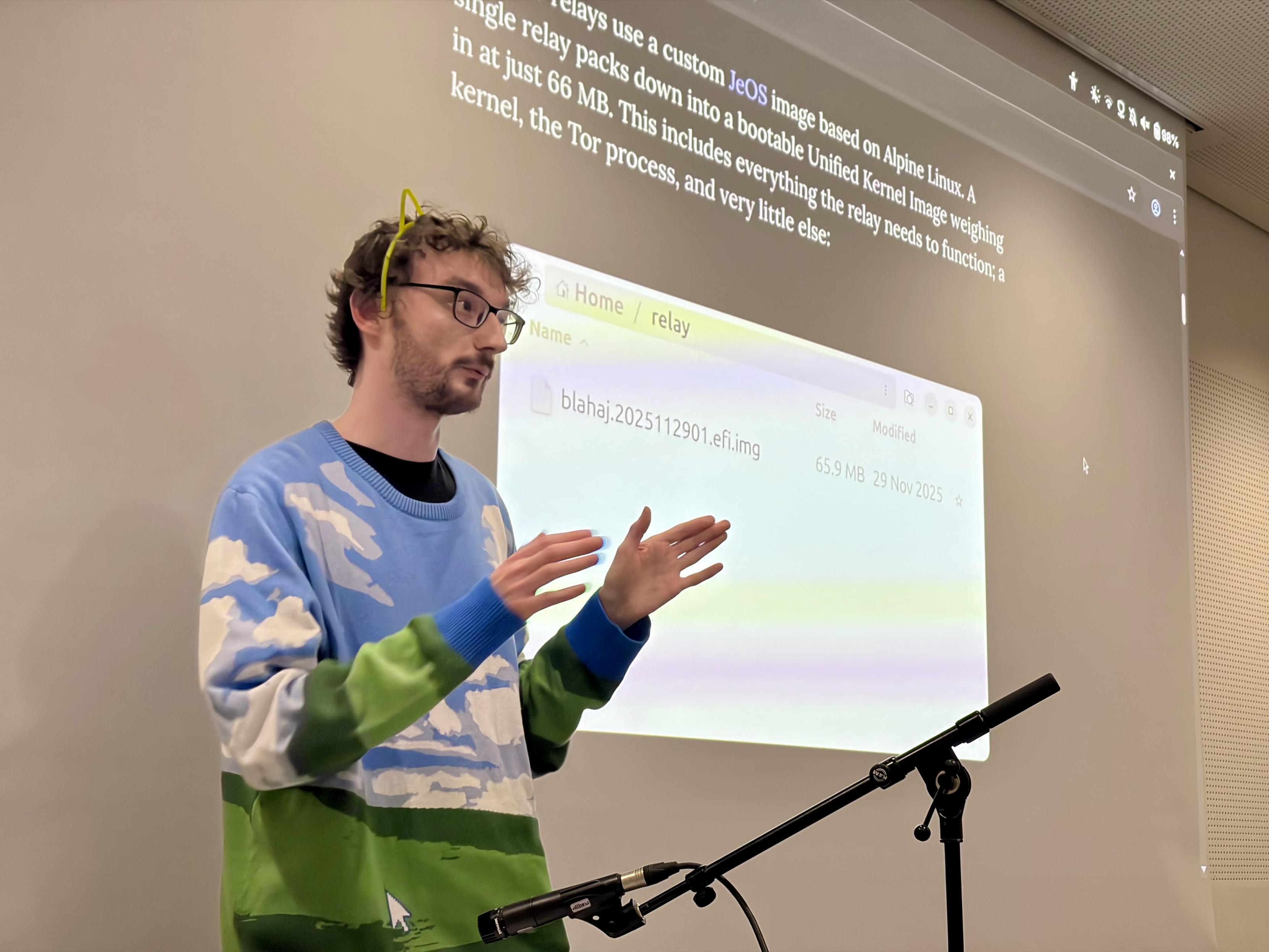 Our volunteer Alex, wearing cat ears and a Windows XP desktop wallpaper sweater, standing in front of a projected screen showing an Emerald Onion blog post.