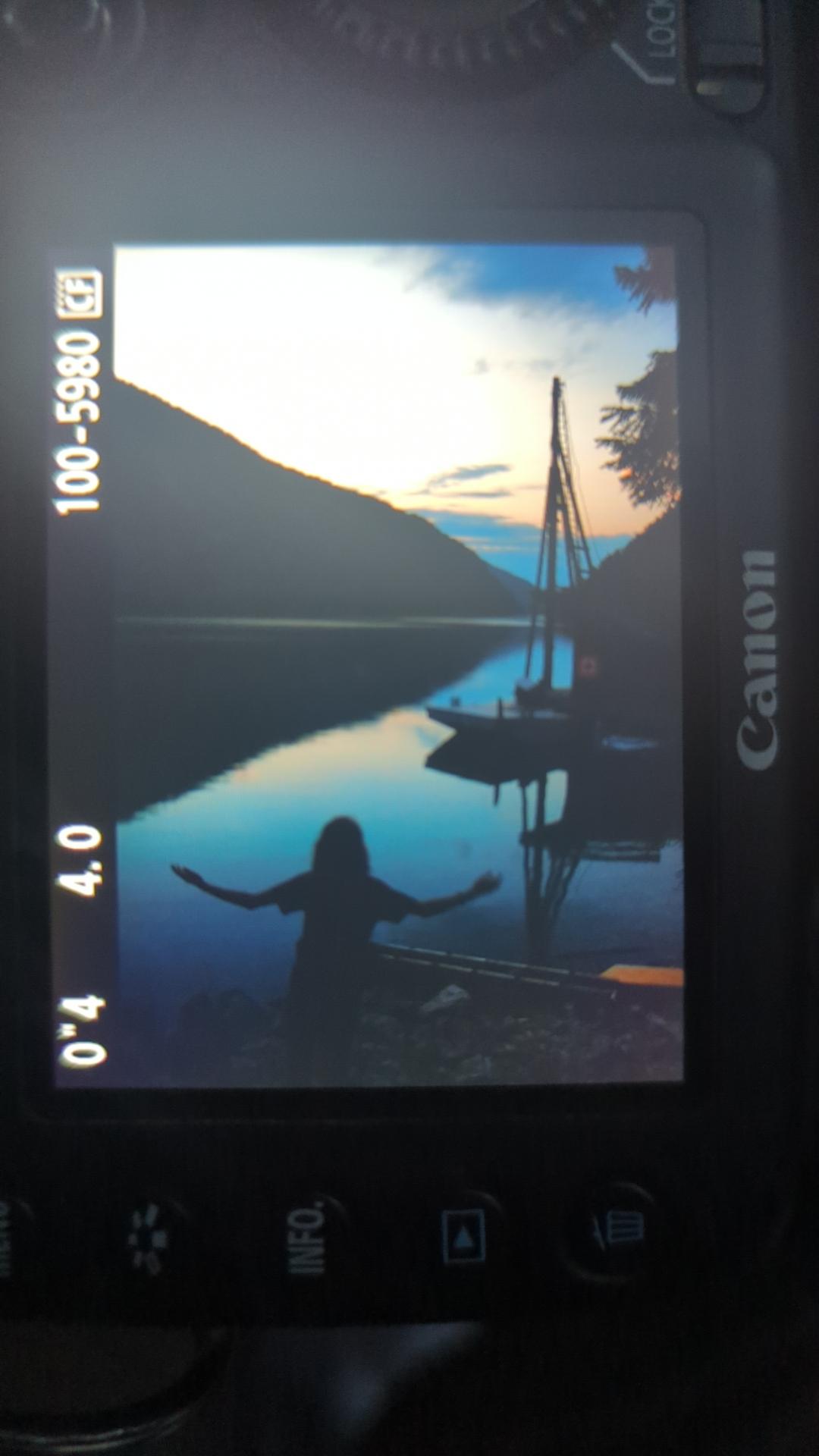 A person (meee!) with their arms open, standing before a lake with a large mountain in the background and pontoon/platform to their right. The sky is orangy from the sunset and the mountains and platform are fully reflected in the slightly blurred water.