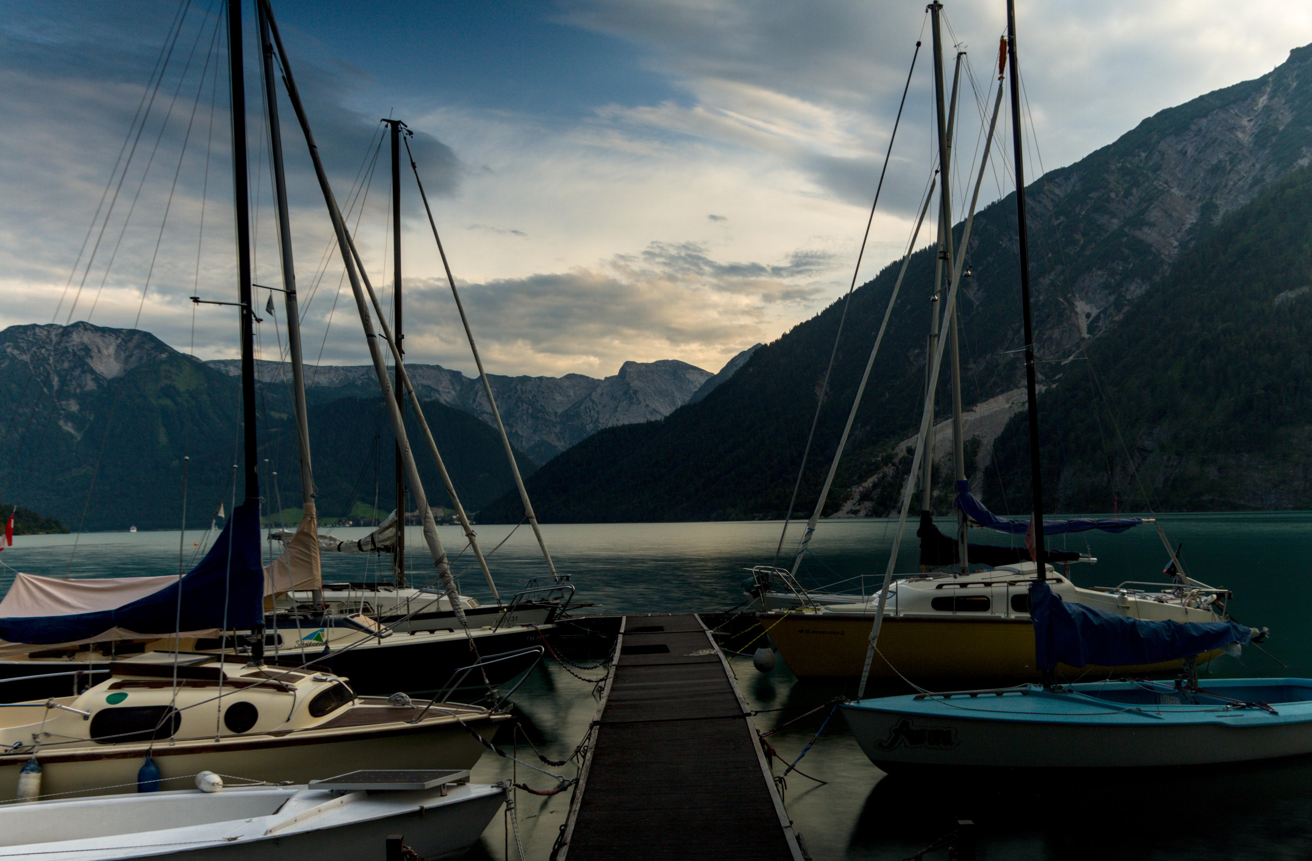 6ish small yachts (maybe 3m long), with their masts not fully in frame, moored to a pontoon in facing pairs, there is massive mountains bordering the lake, which has blurred water.